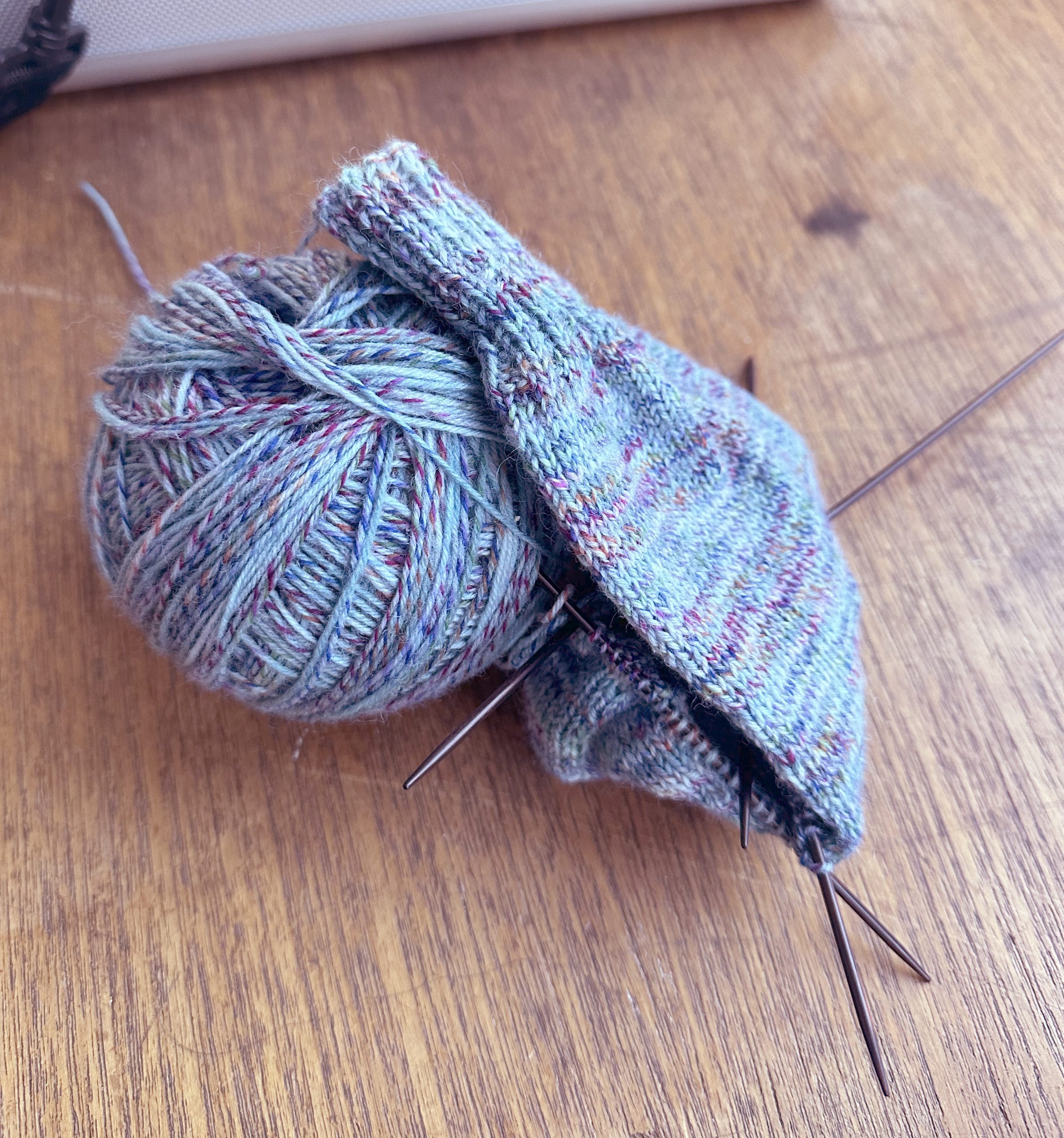 Ein Wollknäuel mit Stricknadeln liegt auf einer Holzfläche.